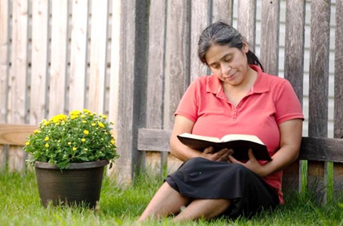 Una fotografía de una dama latina sentada en la grama de un patio, un florero a su lado y una verja de tablas tras ella, en el acto de leer la Biblia.