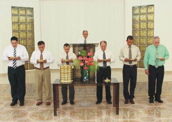 Una fotografía de siete varones al frente de una congregación de la Iglesia de Cristo en el acto de dirigir y repartir la Cena del Señor, todos vestidos de camisa de vestir con corbata, parados sobre un piso de grandes lozas de diseño dorado y tras ellos una pared blanca con paneles de bloques de vidrio dorado en cada lado y sosteniendo platos con el pan de levadura mientras oran.