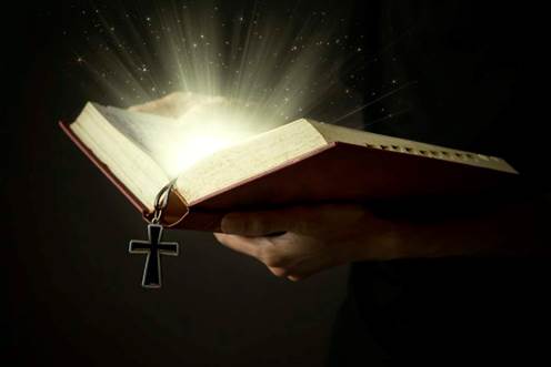 Imagen de una Biblia abierta sostenida en manos varoniles de la cual sube un resplandor blanco, todo contra un trasfondo negro.