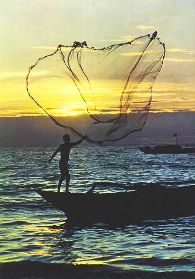 Una fotografía de un varón parado en la proa de un barco pequeño de pesca en el acto de tirar por el aire una red grande que se abre delante de él antes de caer al agua, al atardecer bajo cielos teñidos de amarillo claro sobre tierras ya en tinieblas al otro lado de las aguas.